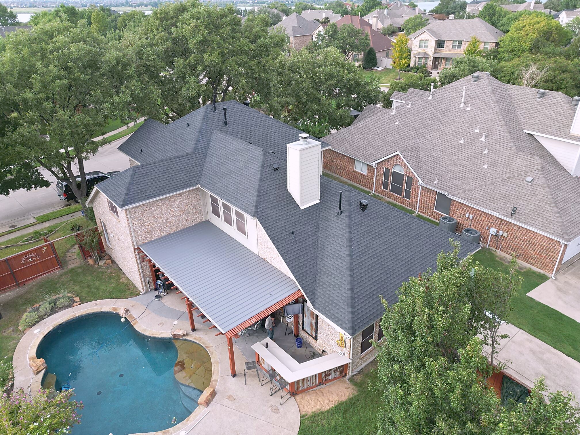 Aerial view of a finished dark shingle roof and a gray metal patio cover on a two-story brick home beside a swimming pool