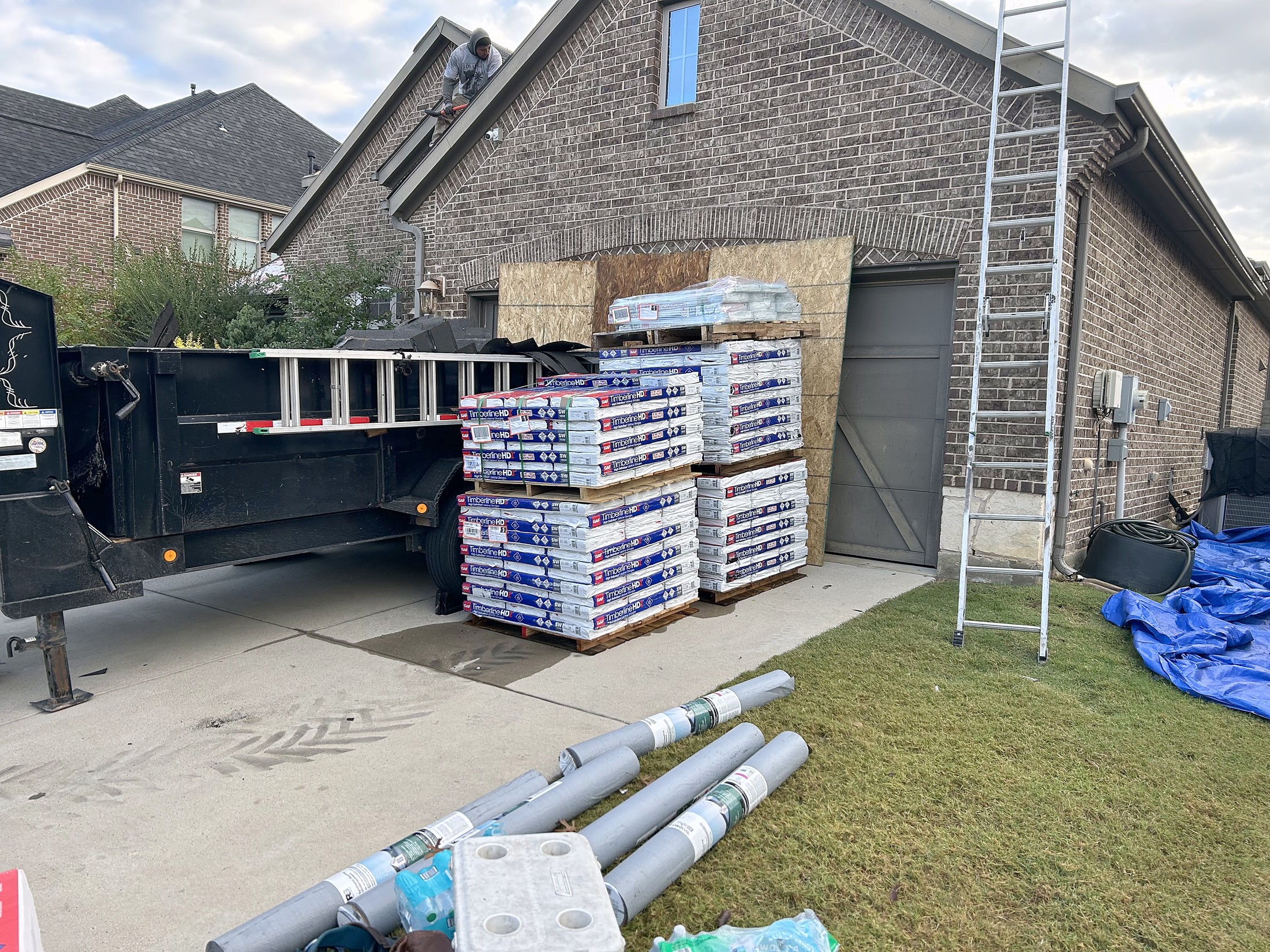 Pallets of shingles and rolls of underlayment staged in a driveway beside a dump trailer